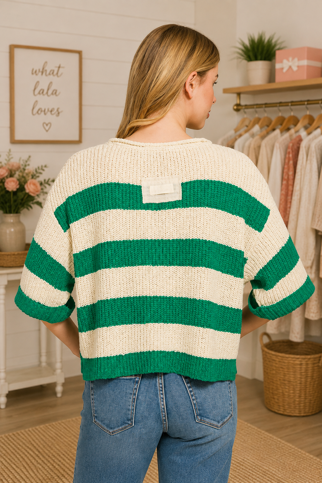 A woman viewed from behind wearing a cream and green striped knit sweater with elbow-length sleeves, paired with blue jeans in a boutique setting.