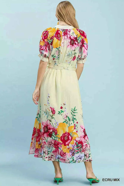 Woman wearing an Umgee ecru floral border print midi dress with a waist tie, puff sleeves, and colorful floral details. Back View