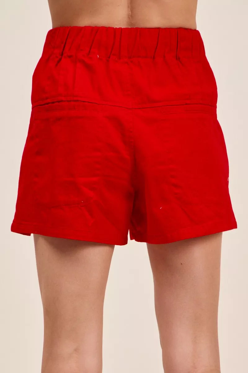 Woman wearing red high waist shorts with pintuck detailing, button-front accents, elastic waistband, and adjustable waist tie. Back View