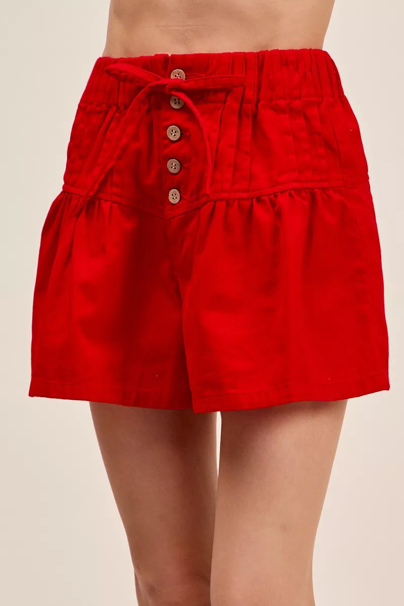 Woman wearing red high waist shorts with pintuck detailing, button-front accents, elastic waistband, and adjustable waist tie. Front View