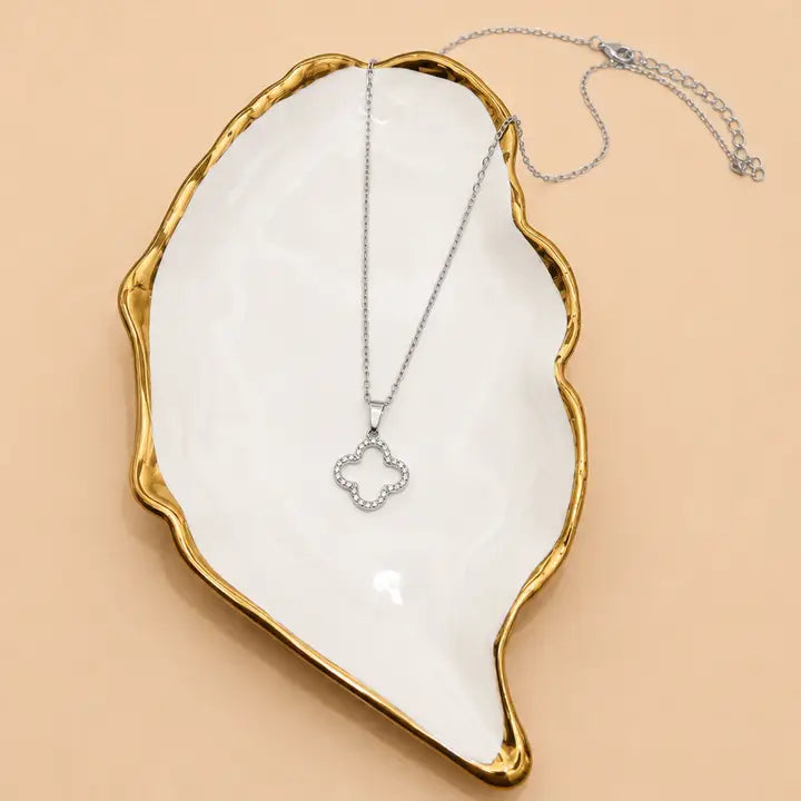 A silver-toned necklace with a small clover-shaped pendant set with clear stones, displayed on a white ceramic dish with a gold rim against a warm beige background.