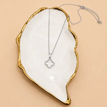 A silver-toned necklace with a small clover-shaped pendant set with clear stones, displayed on a white ceramic dish with a gold rim against a warm beige background.